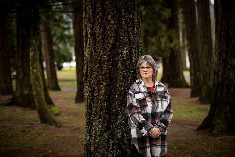 A woman with shoulder-length grey hair poses for a photo against a Douglas fir tree.