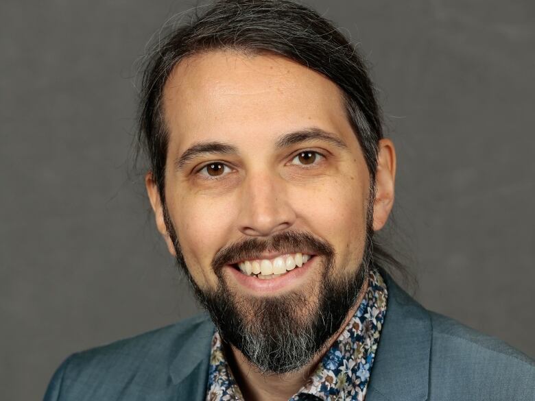 Raiche has a beard and is wearing a blue suit and flowered shirt and is smiling in this headshot