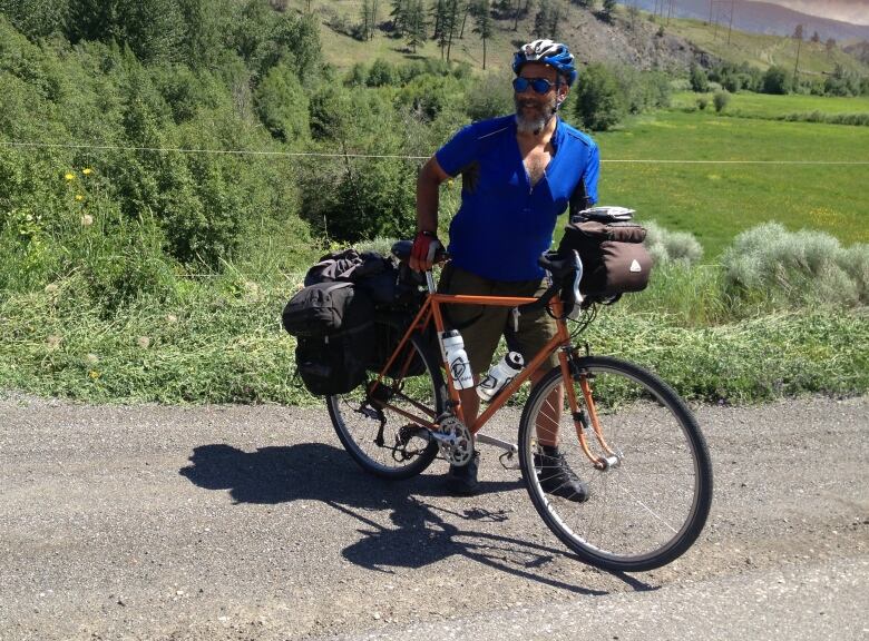 A man in a blue biking outfit and shirt is holding an orange bike in front of a grassy hill. 