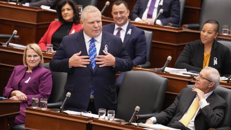 Doug Ford stands in the Legislature, with several members of his caucus seated around him.  