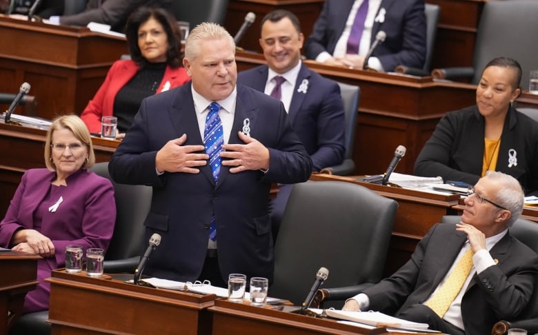 Doug Ford stands in the Legislature, with several members of his caucus seated around him.  