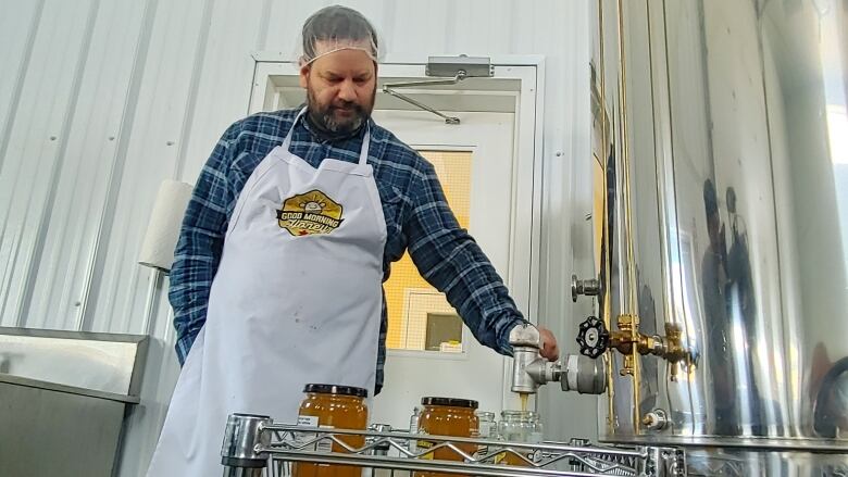 A white man with dark hair and a thick black beard, with patches of grey, has his head covered by a hairnet. He is wearing an apron over his blue long-sleeve flannel shirt. Inside a warehouse, his left hand pushes the lever of an aluminum container, allowing honey to flow into a glass jar.