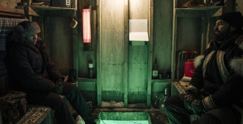 Two people sit across each other while sitting in an ice shack, both wearing warm winter clothing.