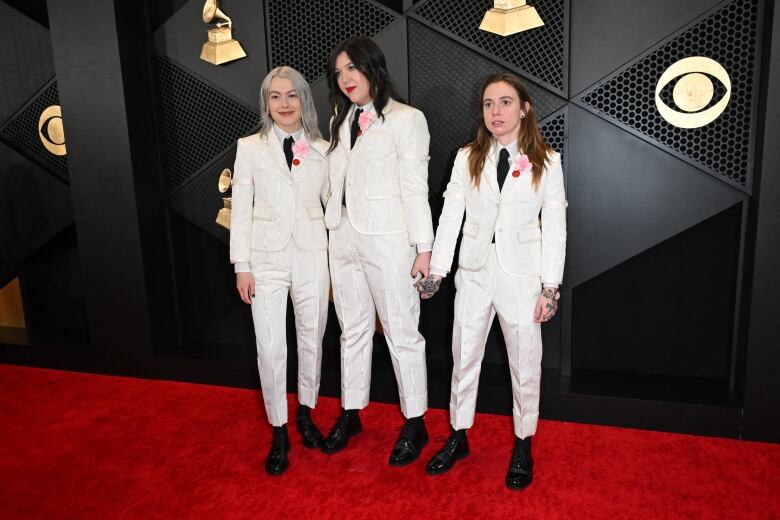 Three women wearing matching white suits with black shoes and ties stand together on a red carpet.