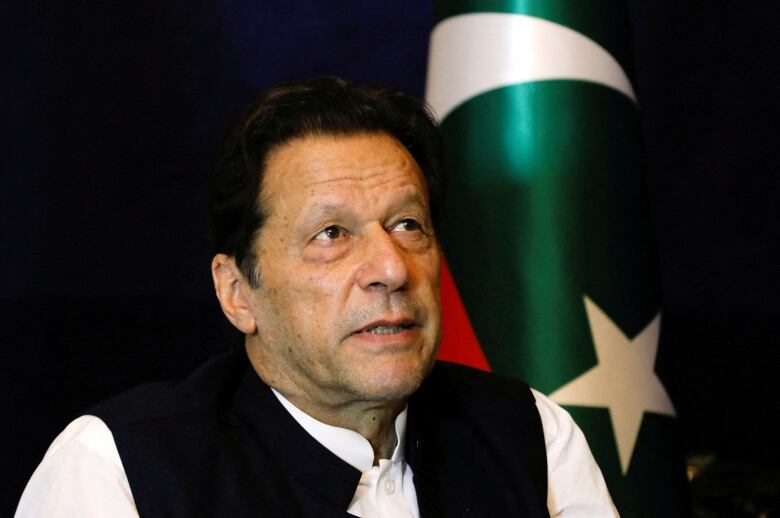 A man seated in front of Pakistan's flag looks upward.