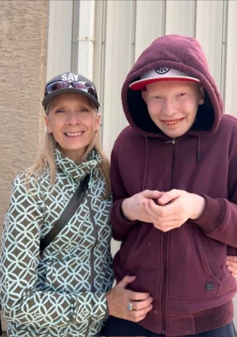 A blond woman puts her arm around a young man wearing a maroon hoodie and ball cap.