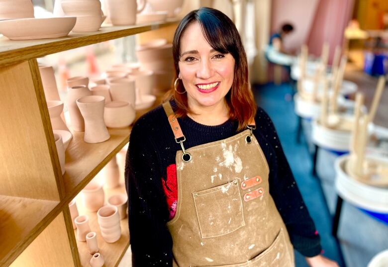 A woman with brown hair wearing an apron is shown leaning against a shelf filled with ceramic vases and containers. Behind her are pottery wheels.