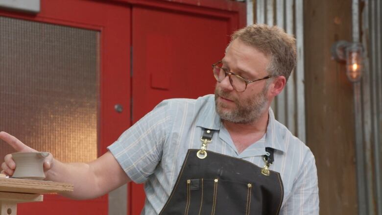 Seth Rogen is shown wearing an apron holding a newly made ash-tray out of clay. He had a beard and tortoise-coloured glasses.