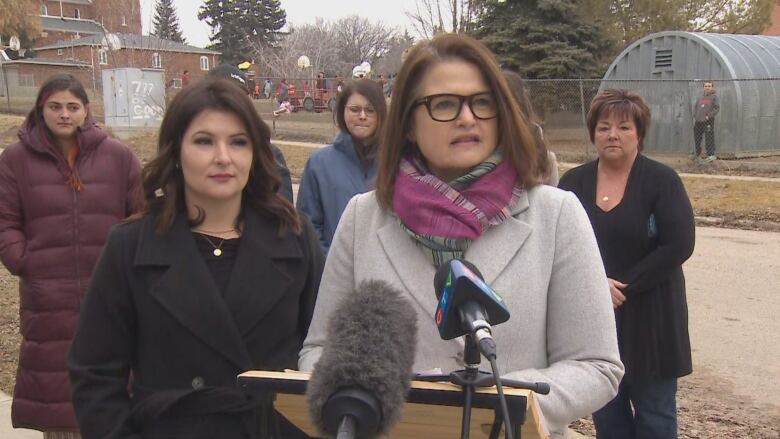 Two women, the one on the left wearing a dark coat and the one on the right wearing a white coat, stand in front of microphones, with other adults and a school playground in the background.