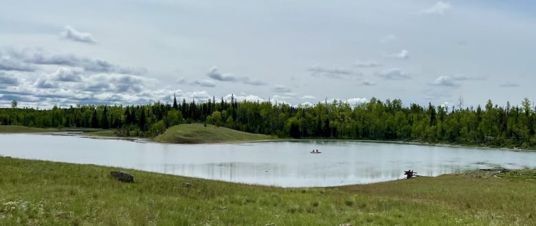 A shallow lake is pictured among greenery. A small boat with two people is in the middle. 