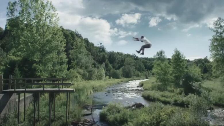 A man in a white shirt and dress pants is depicted flying across a river valley toward a broken down wooden bridge.