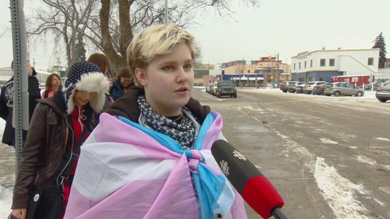 A teenager with short blonde hair stands near a snowy street. He is wrapped in a trans pride flag of pink, white and blue. 