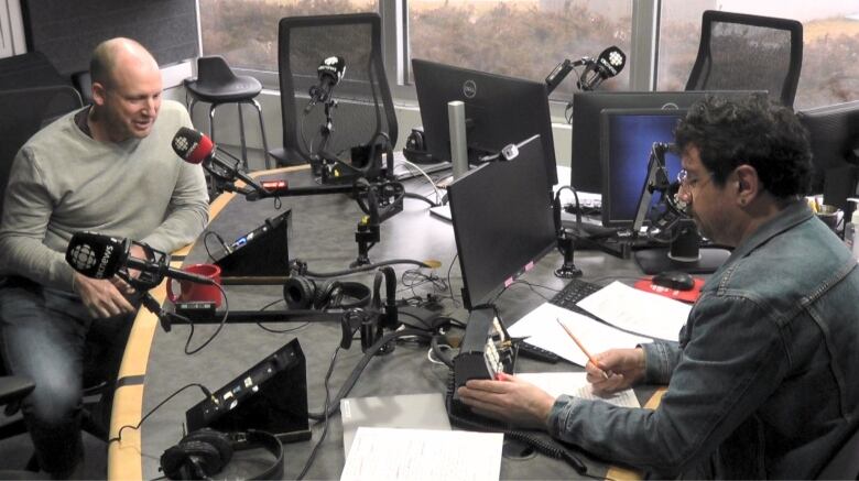 two people sit in a room with audio recording equipment. 