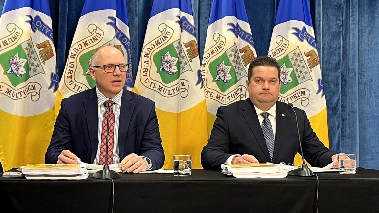 Two men sit at a table with microphones and stacks of paper in front of them. The man on the left is speaking. There are blue and yellow flags on flagpoles behind them.