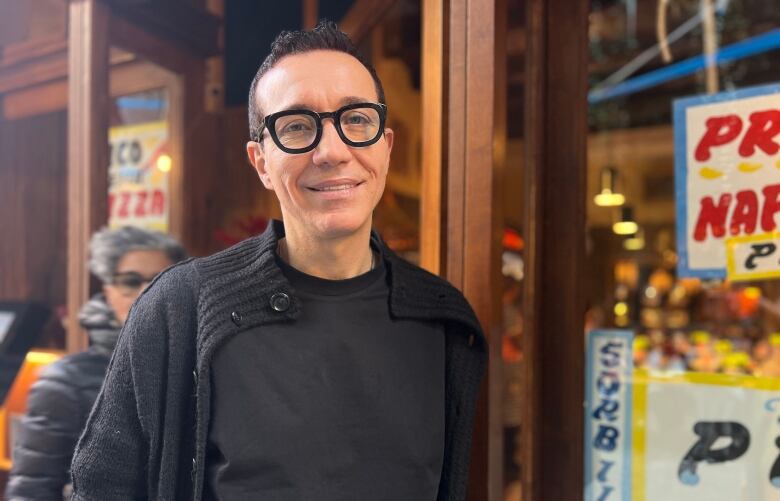 A man with short, dark hair wearing black clothing and black-framed glasses stands in front of a restaurant. 