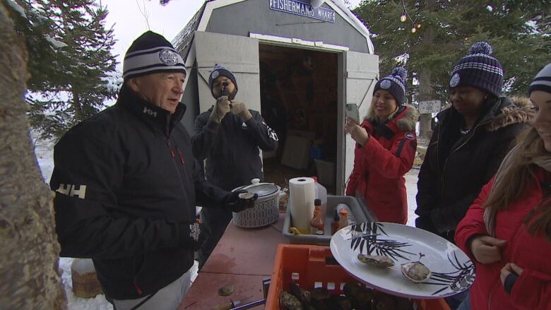 A man in a blue jacket and toque talks to a group in winter jackets about oysters. 