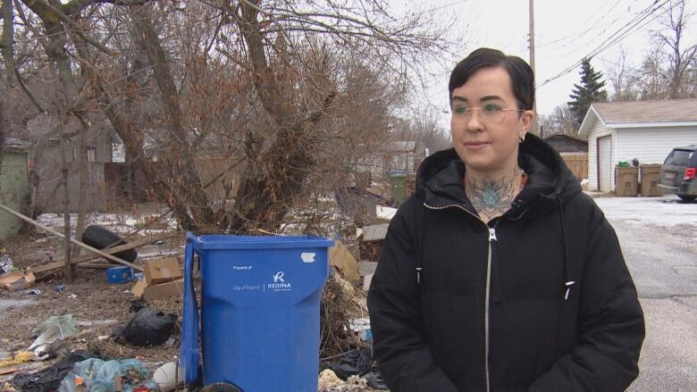 A woman stands in a back alley, with a blue bin surrounded by garbage behind her.