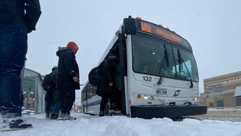 A low-angle shot shows a thick layer of snow on the ground as people step onto a waiting bus.