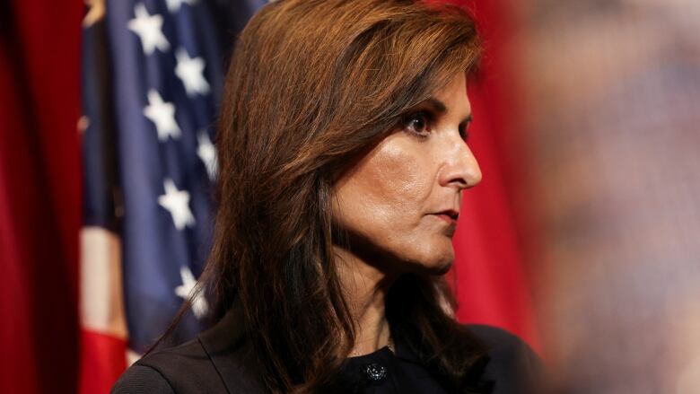 A woman with a serious expression shown from side view with an American flag in the background.