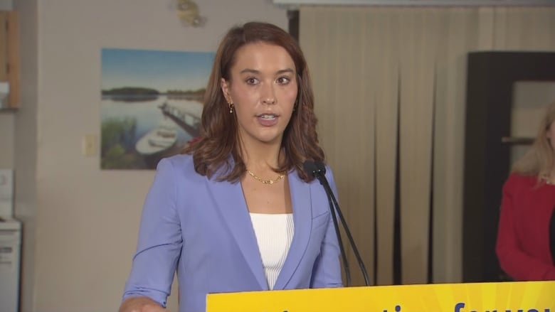 A woman with brown hair in a suit jacket stands at a microphone.