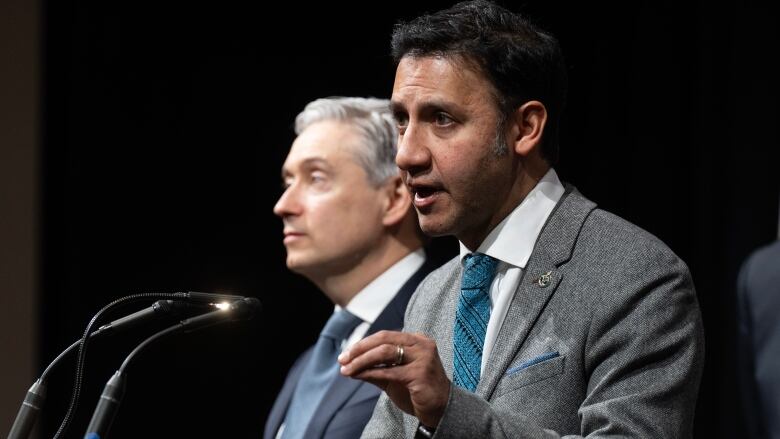 A man in a grey suit gestures with his hand as he speaks at a podium. Another man in a blue suit stands beside him.