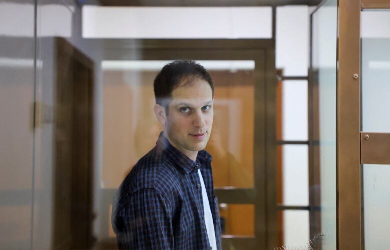 A man in a light and dark blue patterned dress shirt looks directly at a camera through a plexiglass panel in a courtroom holding cell. 