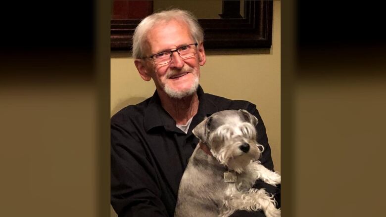 A man with short grey hair and glasses wearing a black long-sleeve shirt holds a grey-haired dog on his lap.