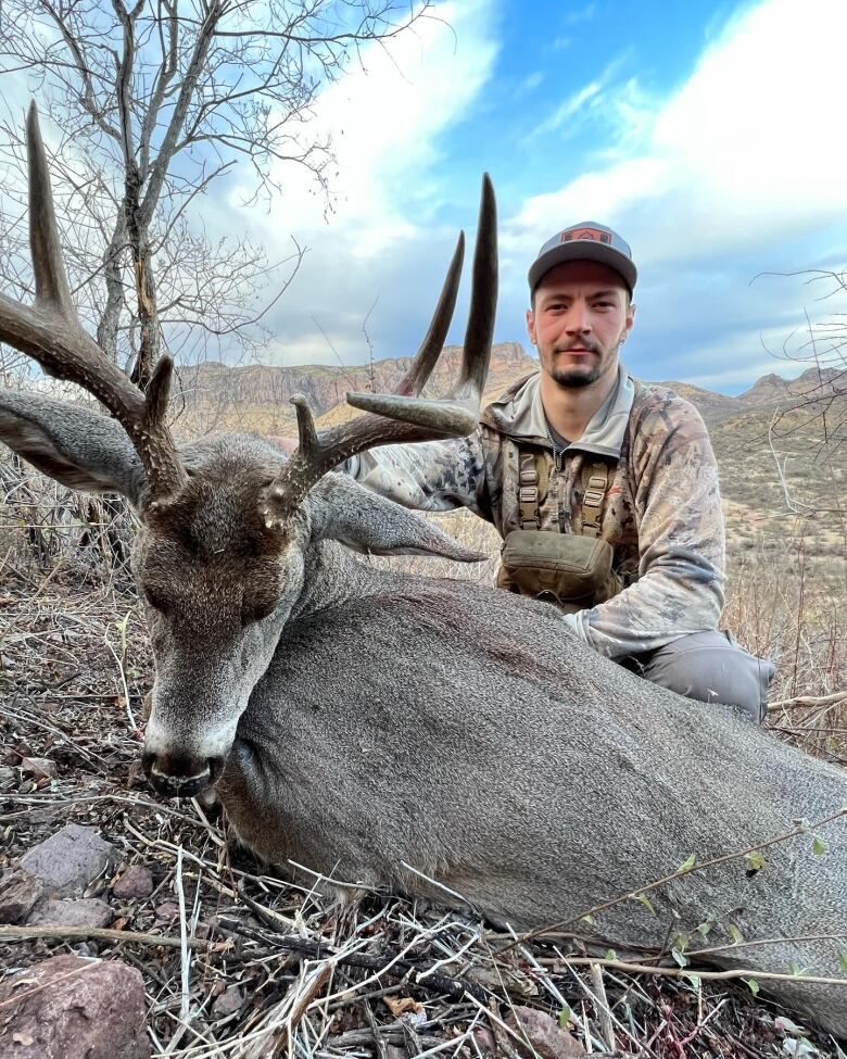 A man sits next to a dead elk holding its antlers up with a mountain range behind.