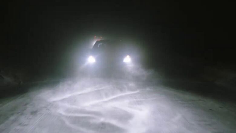 It's night time. A car with bright white headlights drives down an isolated, snowy highway. It is followed by a semi-truck.