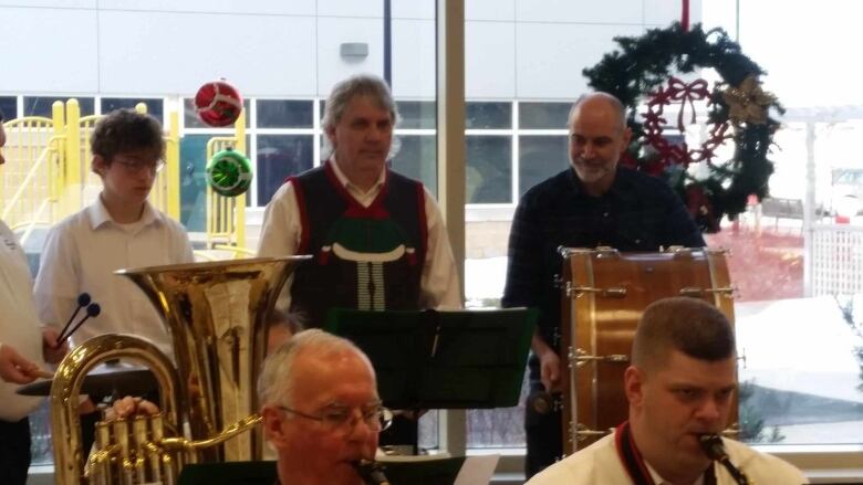 A boy and two men play percussion instruments in a concert band, with a green Christmas wreath to their right. 