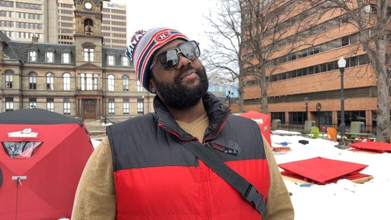 A man in sunglasses and a toque, with a beard, stands in front of Halifax City Hall.