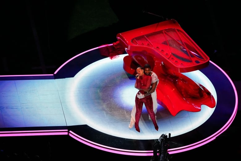 A man in a white suit embraces a singing woman in a red dress in front of a piano  on a  stage