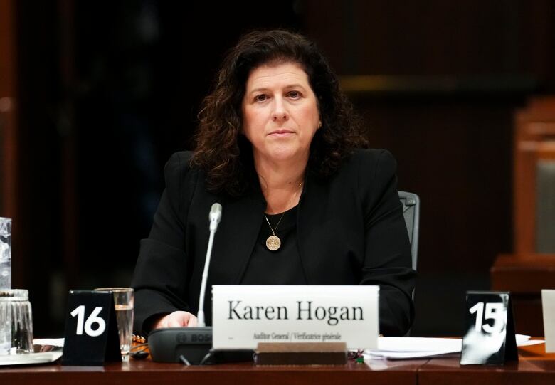 A woman in a black blazer and black shirt sits at a table in front of a microphone.