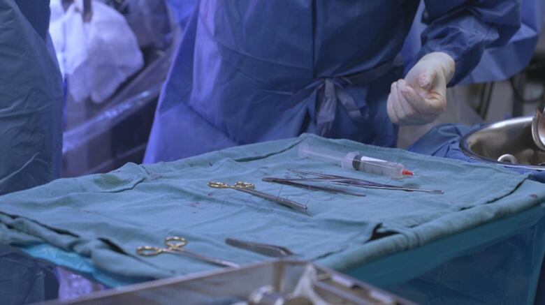 A surgeon's gloved hand reaches toward surgical instruments on a table in a hospital operating room. 