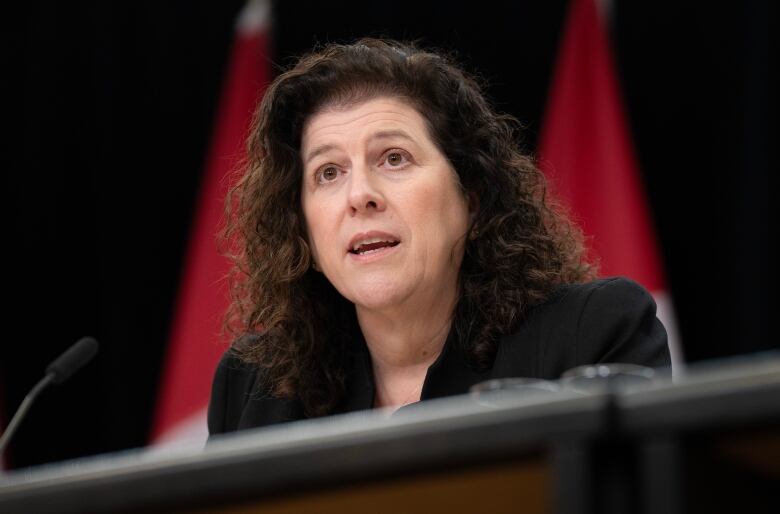 A woman with long, curly brown hair speaks at a microphone with Canadian flags behind her. 