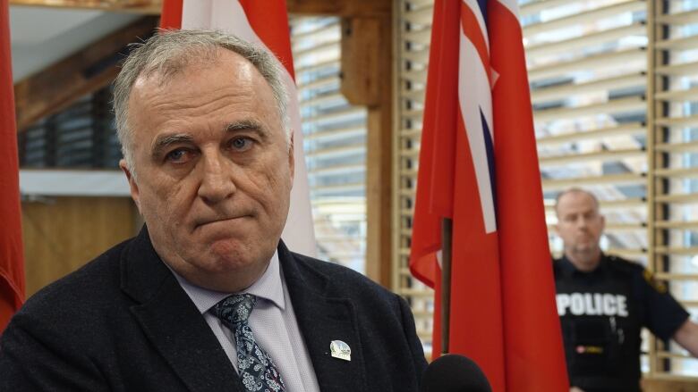 An older man with thinning white hair has a sad look on his face. Behind him are the flags for Ontario and Canada and a police officer.