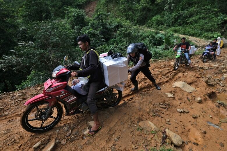 A man in a police uniform pushes a motorcycle, on which sits a man and a box, up a hill.