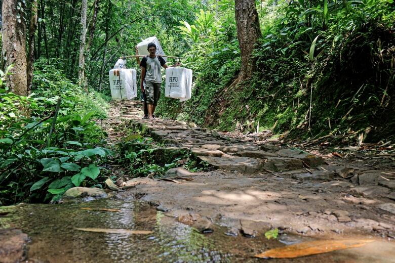 A man carries polling poxes while walking along a creek.