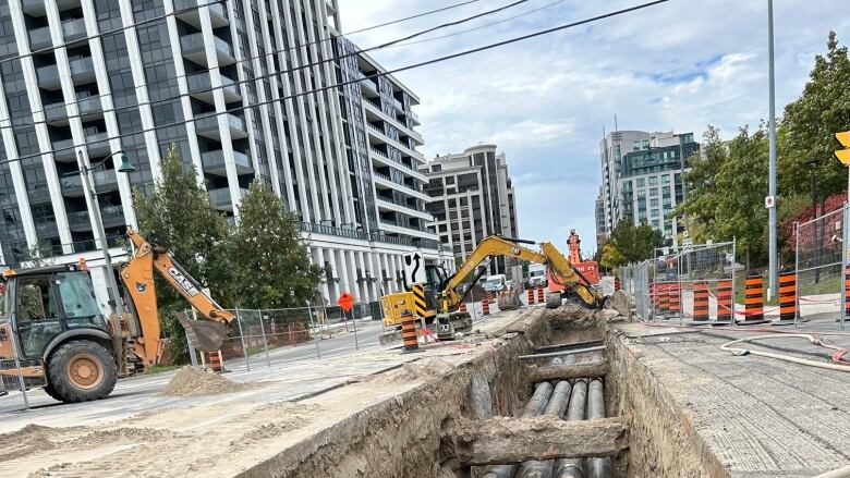 Pipes in a trench run toward and away from the viewer, with excavators and pylons in the distance and highrise buildings on either side.