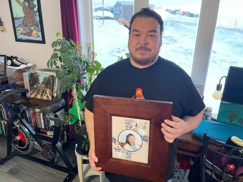 A man stands in a home holding up a framed photo of an elderly woman.