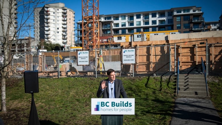 A tall white man stands at a microphone in front of a housing construction site.