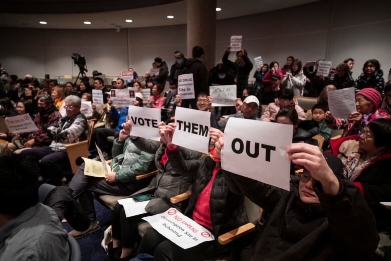 Protesters hold up signs reading 'Vote Them Out' in a packed city hall.