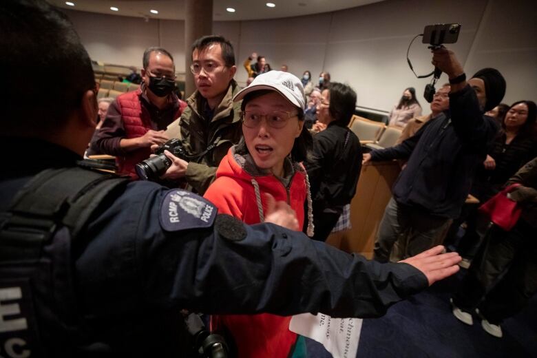 An East Asian woman is escorted out of a council chamber by a uniformed police officer, with other photographers visible.