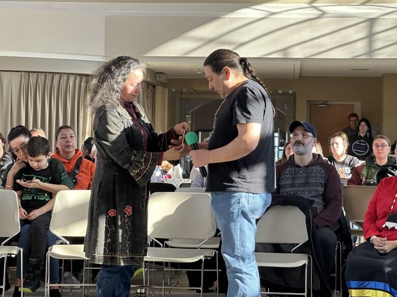 A woman with long, grey hair hands a white string to a man with a long, black braid and black t-shirt.