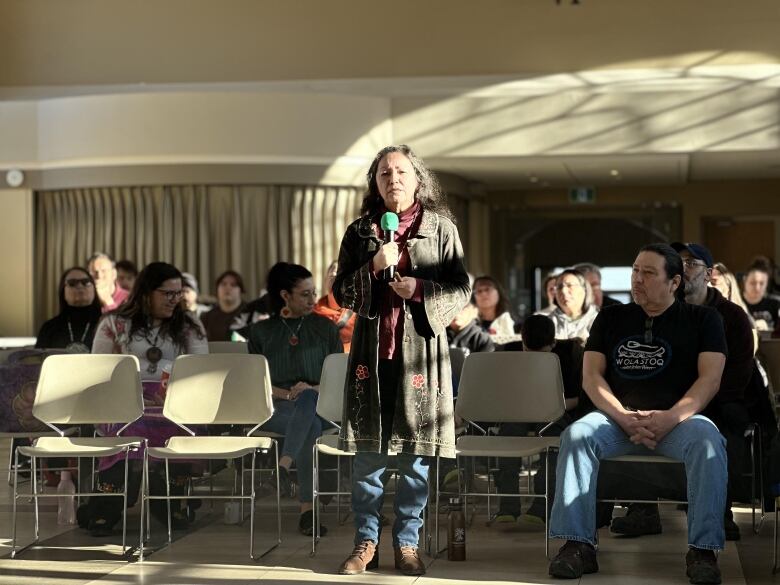 A woman wearing a long, grey cardigan with embroidered flowers stands in a sunbeam, holding a microphone. Lines of people sit behind her.