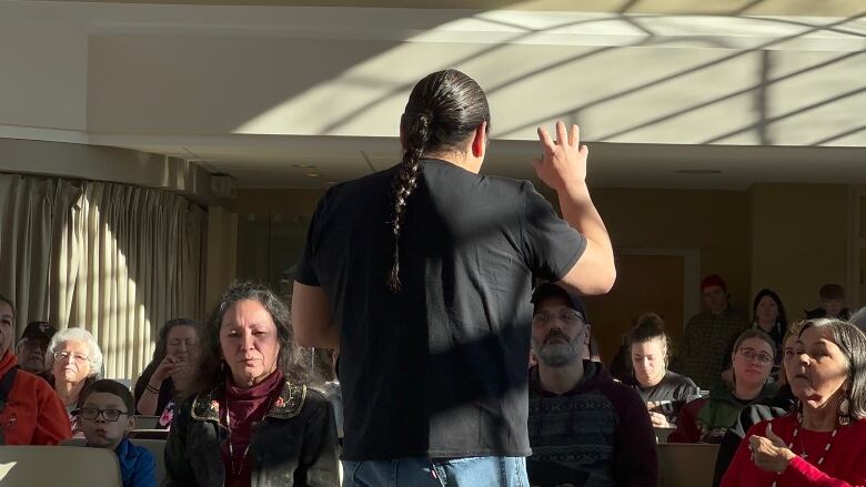 A man wearing a black t-shirt and brown hair braided down his back stands facing away from the camera, gesturing to a congregation. 