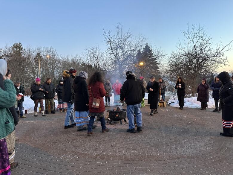 A crowd of people dressed in winter coats stand in a circle around an outdoor fire. There are trees and snow in the distance.