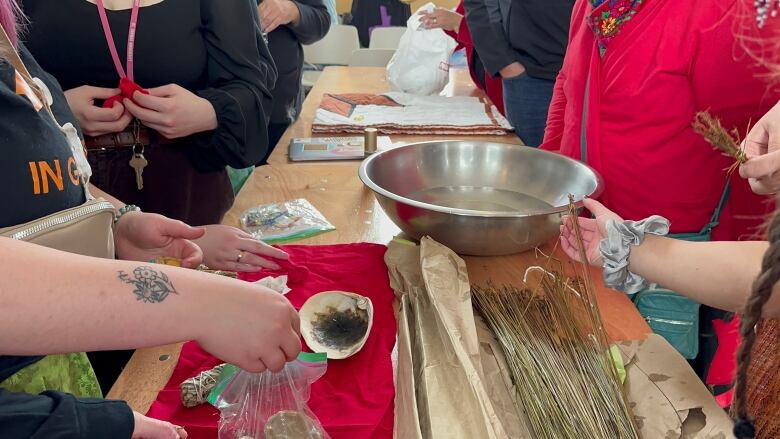 A wooden table shows an assortment of materials, including a bowl full of water and a pile of grass. People stand around the table.