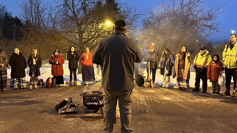 A man stands outside with his back to the camera. He faces a fire and circle of people. 