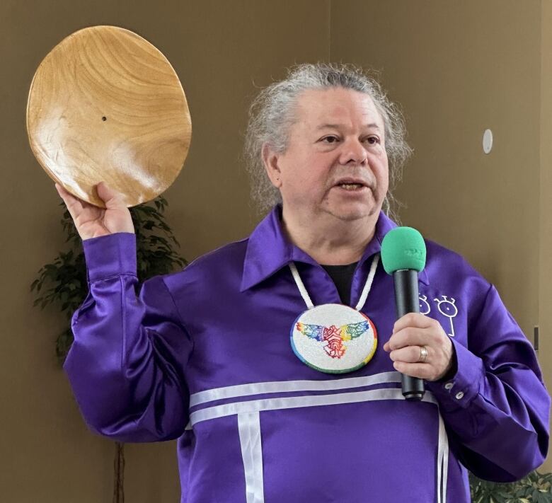 A man with white hair pulled back in a braid holds up a wooden plate.